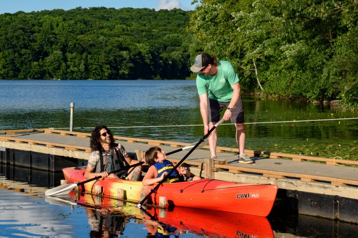 a man and a child sitting in a double kayak as someone standing on the dock hands them paddles